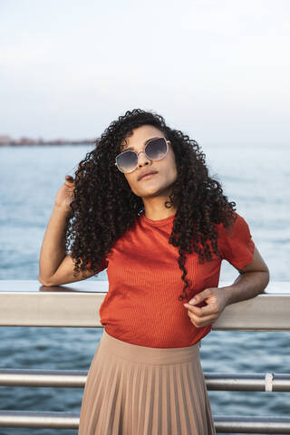 Schöne Frau mit Sonnenbrille, die sich am Meer an ein Geländer lehnt, lizenzfreies Stockfoto
