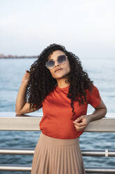 Schöne Frau mit Sonnenbrille, die sich am Meer an ein Geländer lehnt - RCPF00041
