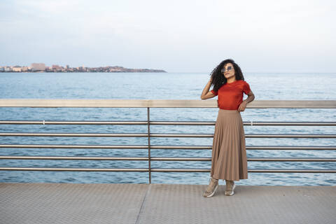 Schöne Frau mit Sonnenbrille, die sich am Meer an ein Geländer lehnt, lizenzfreies Stockfoto