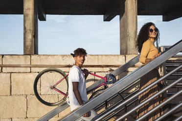 Junges Paar steigt eine Treppe hinauf, trägt ein Fahrrad - RCPF00062