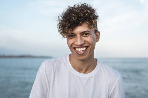 Portrait of laughing young man with curly hair by the sea - RCPF00064