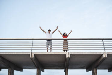 Junges Paar steht auf einer Brücke und winkt mit erhobenen Armen - RCPF00072