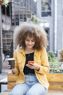 Portrait of smiling teenage girl sitting on bench with coffee to go looking at cell phone - VPIF01533