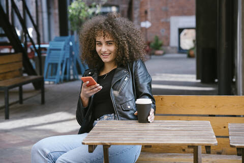 Porträt eines lächelnden Teenagers auf einer Bank sitzend, mit Kaffee zum Mitnehmen und Mobiltelefon, lizenzfreies Stockfoto