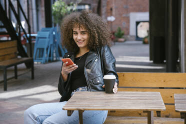 Porträt eines lächelnden Teenagers auf einer Bank sitzend, mit Kaffee zum Mitnehmen und Mobiltelefon - VPIF01540