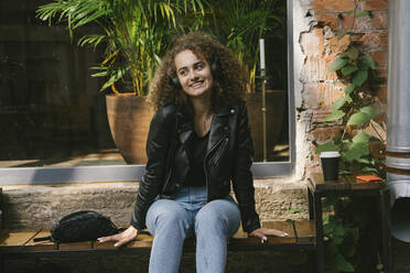 Porträt eines lächelnden Teenager-Mädchens, das in einem Café im Freien sitzt und mit Kopfhörern und Smartphone Musik hört - VPIF01548