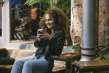 Portrait of smiling teenage girl sitting at outdoor cafe with coffee to go listening music with headphones - VPIF01549