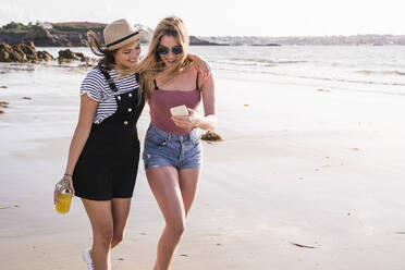 Two girlfriends having fun, walking on the beach, taking smartphone selfies - UUF19029