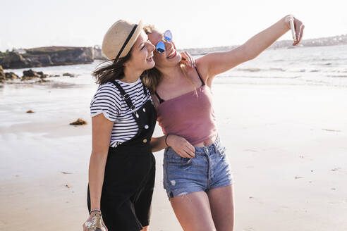 Two girlfriends having fun, walking on the beach, taking smartphone selfies - UUF19031