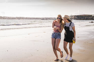 Two girlfriends having fun, walking on the beach, taking smartphone selfies - UUF19035