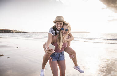 Zwei Freundinnen haben Spaß am Strand, tragen sich gegenseitig Huckepack und machen Smartphone-Selfies - UUF19044