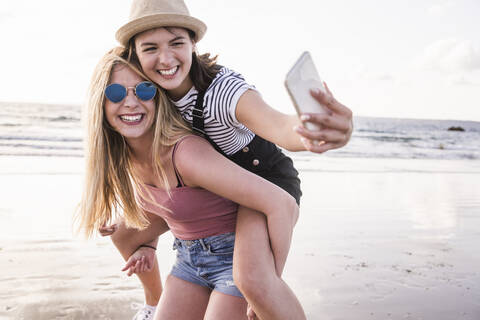 Zwei Freundinnen haben Spaß am Strand, tragen sich gegenseitig Huckepack und machen Smartphone-Selfies, lizenzfreies Stockfoto