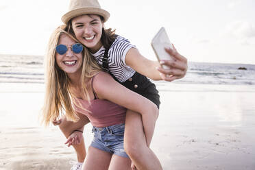 Zwei Freundinnen haben Spaß am Strand, tragen sich gegenseitig Huckepack und machen Smartphone-Selfies - UUF19045