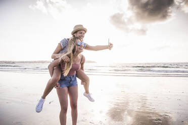 Zwei Freundinnen haben Spaß am Strand, tragen sich gegenseitig Huckepack und machen Smartphone-Selfies - UUF19046