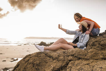 Two girlfriends sitting on rocky beach, taking smartphone selfies - UUF19047