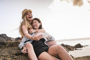Two girlfriends sitting on rocky beach, taking smartphone selfies - UUF19049