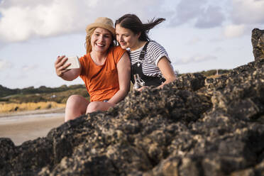 Two girlfriends sitting on rocky beach, taking smartphone selfies - UUF19054