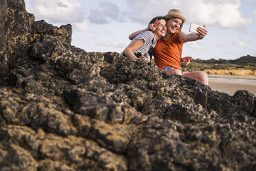 Zwei Freundinnen sitzen am felsigen Strand und machen Smartphone-Selfies - UUF19055