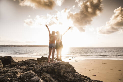 Zwei Freundinnen stehen am felsigen Strand, winken bei Sonnenuntergang, Rückansicht - UUF19056