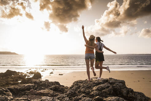 Zwei Freundinnen stehen am felsigen Strand, winken bei Sonnenuntergang, Rückansicht - UUF19058