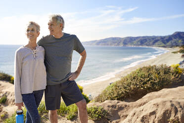 Lächelndes älteres Paar, das auf einer Klippe am Strand steht, gegen den Himmel an einem sonnigen Tag - CAVF64940