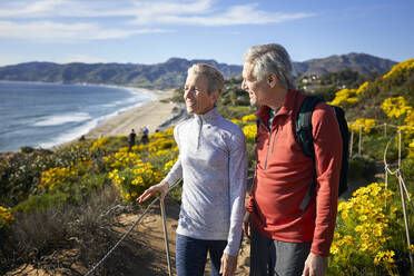 Lächelndes älteres Paar, das sich bei einem Spaziergang auf einem Berg am Strand gegen den Sommerhimmel unterhält - CAVF64941
