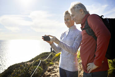 Lächelndes älteres Paar, das ein Selfie macht, während es am Meer gegen den Himmel steht, während eines sonnigen Tages - CAVF64944