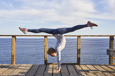 Full length senior woman doing handstand while exercising on floorboard by sea against sky - CAVF64949