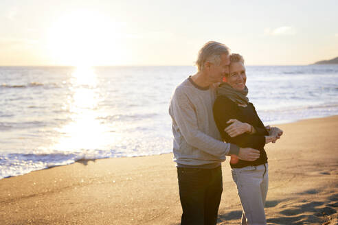 Zärtliches älteres Paar steht am Strand gegen den Himmel bei Sonnenuntergang - CAVF64963