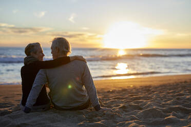 Rear view of senior couple talking while sitting on sand at beach during sunset - CAVF64965