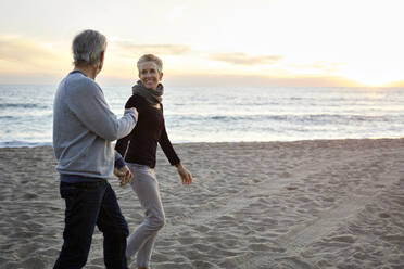 Smiling senior couple holding hands while walking at beach during sunset - CAVF64967