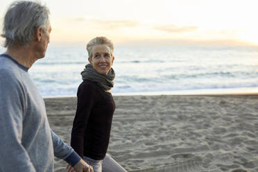 Seitenansicht eines älteren Paares, das sich bei einem Spaziergang am Strand während des Sonnenuntergangs an den Händen hält - CAVF64968