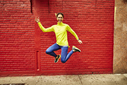 Eine Frau in sportlicher Kleidung springt vor einer leuchtend roten Wand. - CAVF65018