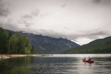 Mann und Kind paddeln mit einem roten Kanu auf einem Bergsee an einem bewölkten Tag - CAVF65037
