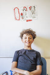 Porträt eines lächelnden Jungen, der vor einer Zeichnung auf einem Whiteboard sitzt - DLTSF00221