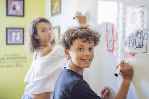 Porträt eines lächelnden Jungen, der auf einem Whiteboard mit einem Mädchen im Hintergrund zeichnet - DLTSF00223