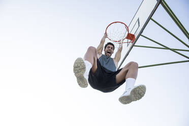 Young man screaming while hanging on basketball hoop and looking at camera - ABZF02627