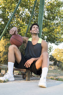 Junger Sportler sitzt auf einem Sportplatz, hält einen Basketball und lacht, während er wegschaut - ABZF02628