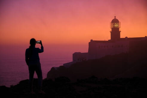 Portugal, Algarve, Silhouette einer Person, die Smartphone-Fotos von Cape Saint Vincent Leuchtturm in der Morgendämmerung - MRF02163