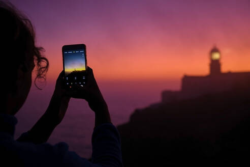 Portugal, Algarve, Blick über die Schulter einer Person, die Smartphone-Fotos von Cape Saint Vincent Leuchtturm in der Morgendämmerung - MRF02165