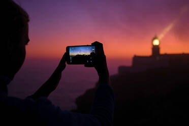 Portugal, Algarve, Blick über die Schulter einer Person, die Smartphone-Fotos von Cape Saint Vincent Leuchtturm in der Morgendämmerung - MRF02166