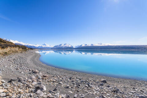 Neuseeland, Südinsel, Klarer Himmel über felsigem Ufer des Lake Pukaki mit Bergen im Hintergrund, lizenzfreies Stockfoto