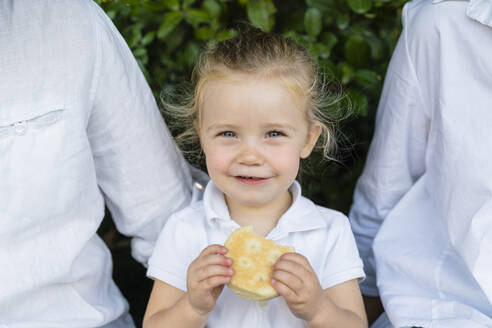 Porträt eines kleinen Mädchens mit einem Snack inmitten der Eltern - MGIF00765