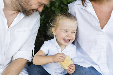Glückliches kleines Mädchen mit einem Snack inmitten der Eltern sitzend - MGIF00766