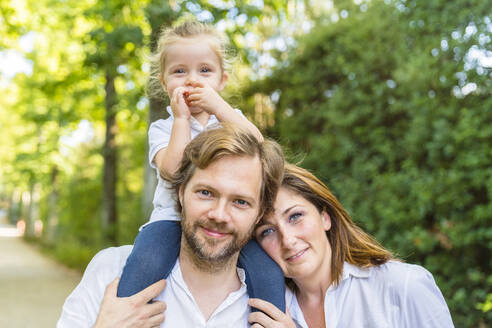 Porträt einer glücklichen Familie in einem Park mit einem Vater, der seine kleine Tochter auf den Schultern trägt - MGIF00773