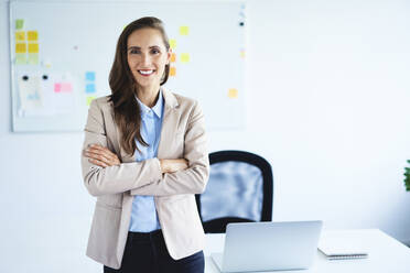 Cheerful young businesswoman looking at camera with crossed arms standing in office - BSZF01475