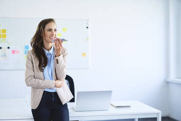 Young woman leaning on desk in office during phone call - BSZF01479