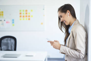 Young businesswoman standing in office using phone and wireless earphones - BSZF01487