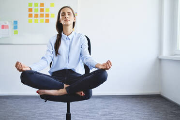 Junge Geschäftsfrau sitzt auf einem Bürostuhl und meditiert - BSZF01505