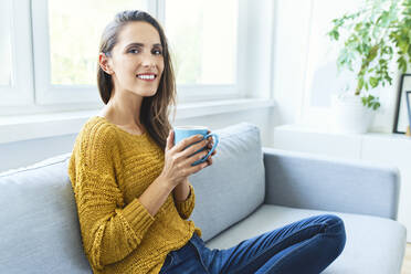 Portrait of young woman drinking coffee sitting on sofa and smiling at camera - BSZF01531